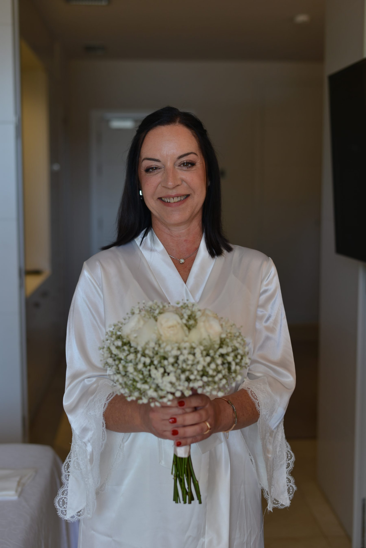 Novia con bata de seda color marfil sujetando un ramo de flores blancas en el pasillo del hotel, con maquillaje natural luminoso